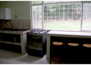 Bright kitchen with a gas stove under a large window, a sink to the left, and a marble counter extending along the window with three wooden bar stools nearby.