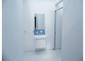 Small white bathroom sink with a blue tile backsplash and wall-mounted cabinet in a bright, empty room with a towel bar on the left and an open doorway on the right.
