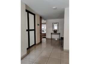 Indoor lobby hallway with frosted glass doors on the left, a brick pillar, and beige tiled flooring; a console table with a mirror and flowers sits toward the far end.