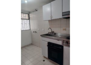 Small white-tiled kitchen with a stainless steel sink, counter, and built-in oven; white upper and lower cabinets and a window with bars on the left side