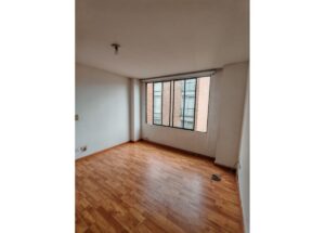 Empty rectangular room with wooden floor, large window with bars, bare ceiling light, and a few cords on the floor.