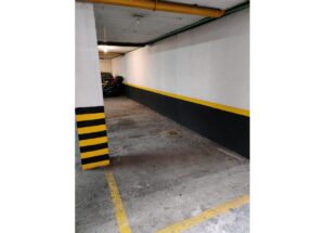 Underground parking area with white walls, a black lower wall stripe, and yellow safety markings; a striped pillar stands at the left and vehicles are parked at the far end.