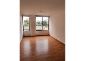 Empty rectangular room with wood laminate flooring and large windows overlooking trees and rooftops outside.