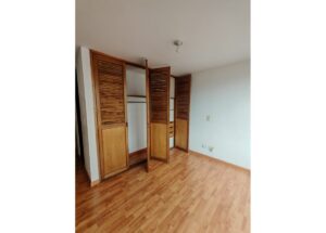 Bedroom with open wooden louvered closet doors, showing shelves and a hanging rod; light wood floor and pale walls.