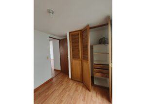 Wooden wardrobe with louvered doors open, revealing shelves and drawers; a bag sits on the top shelf.