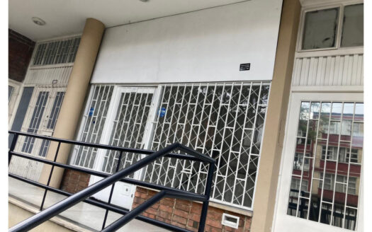 Closed storefront with white metal security grilles across large windows and door; beige columns, brick base, and a black railing along a short ramp outside.
