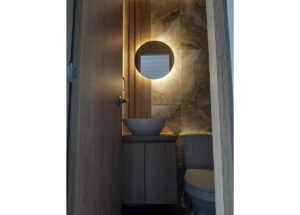 Small modern bathroom with a round backlit mirror above a white vessel sink on a wooden vanity, marble-tiled wall, and a toilet to the right.