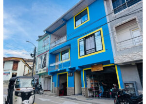 Blue building with yellow window frames on a street; ground-floor shop displays goods, with a tuk-tuk and a motorbike parked nearby.