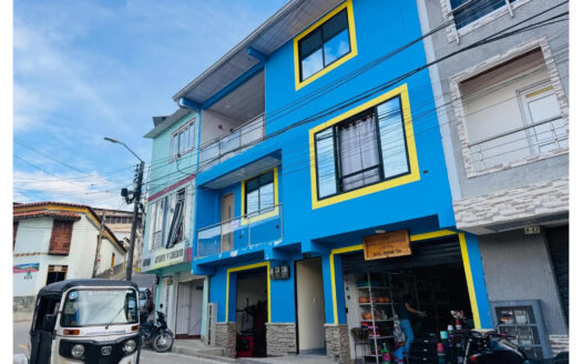 Blue building with yellow window frames on a street; ground-floor shop displays goods, with a tuk-tuk and a motorbike parked nearby.