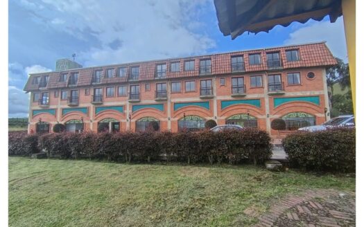 Three-story brick hotel with orange arches on the ground floor, teal storefronts, hedges, and parked cars in front.