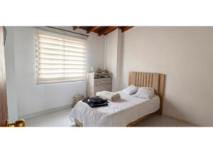 Tidy bedroom with a wooden upholstered headboard, white bedding, and folded towels on the bed; dresser and window with striped blind in soft light.