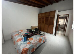 Bedroom with a bed covered by a patterned duvet and clothes piled on top, next to a large wooden wardrobe; open door to a hallway visible in the background.