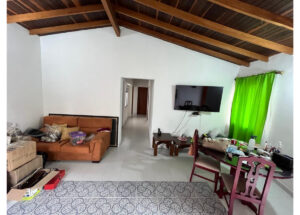 Living room with an orange sofa, clutter on the left, wall-mounted TV, and a bright green curtain on the right under a wooden beam ceiling.