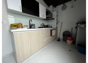 Compact kitchen with light wood cabinets, white countertops, and a black faucet; fruit (pineapple, bananas) on the counter.