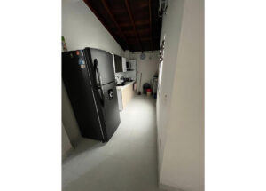 Narrow utility kitchen with a large black refrigerator, white cabinets, and a row of appliances along the counter under an exposed wooden beam ceiling.