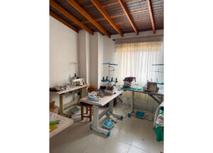 Small sewing workshop with several sewing machines on work tables, spools, and fabric pieces under a wooden ceiling and window light.