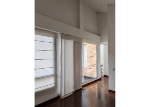 Bright living space with tall windows, white vertical blinds, sheer curtains, and a polished wooden floor.