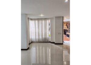 Bright empty living room with glossy white tiles and a large window covered by sheer vertical blinds near an outdoor area.