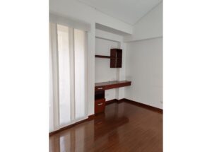Corner of a white room with a wooden wall desk and shelving unit; vertical blinds on the left and polished hardwood floor.