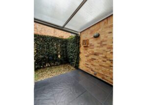 Covered outdoor patio with dark tile floor, brick wall, and green hedges along one side.
