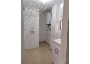 Laundry area with a white foldable drying rack, wall boiler, and a sink cabinet in a white-tiled room.