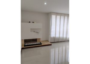 Minimalist white fireplace with a recessed shelf and pebble bed, in a bright living room with sheer white curtains.