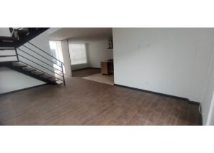 Open-plan living area with a wooden tile floor, white walls, and a metal staircase along the left side; a kitchen is visible in the background.