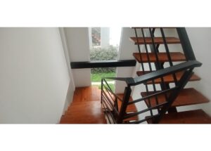 Indoor wooden staircase with black metal railing, viewed from above near a window overlooking a green yard outside the building.