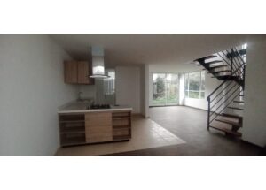 Open-concept kitchen and living area with an island, sink, gas stove, range hood, and wooden cabinets; staircase with metal railing leading upstairs; large windows overlook a garden.