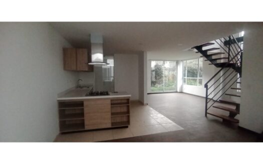Open-concept kitchen and living area with an island, sink, gas stove, range hood, and wooden cabinets; staircase with metal railing leading upstairs; large windows overlook a garden.