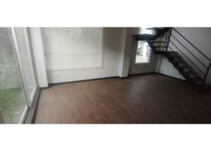 Empty modern living space with dark wood flooring, large windows along the left wall, and a metal staircase on the right brought to the upper level.