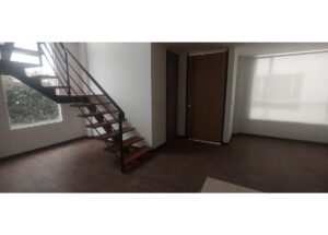 Empty modern living space with a wooden open-riser staircase and metal railing to the left. The room has dark wood flooring and a large window on the far wall.