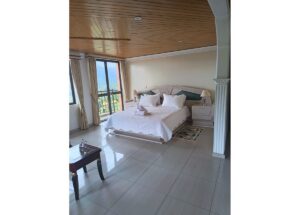 Bright hotel bedroom with a large white bed, two nightstands, and a wood-paneled ceiling with a mountain view through balcony doors.