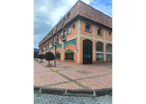 Two-story brick building with arched turquoise storefronts, large windows, and a tiled red roof in a brick-paved plaza with street lamps and trimmed trees, outdoors.