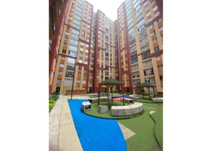 Courtyard between tall, colorful apartment buildings with blue path, green turf, and shaded seating areas.