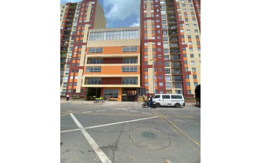 Tall residential building with orange and beige panels; ground-level entrance, white van and motorcycles parked outside on a city street.