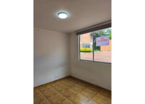 Empty corner of a small room with parquet flooring and a window showing a 'For Sale' sign outside; neutral walls and ceiling light on.