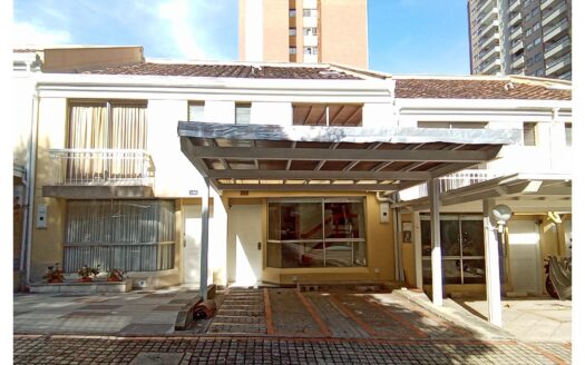 Two-story beige townhouse with a metal-framed carport and large front window, set between neighboring buildings on a sunny day.