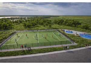A fenced artificial-turf soccer field with players, spectators along the sideline, and open green landscape beyond.