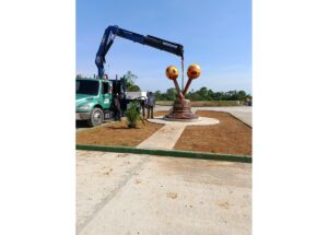 Crane truck installs a public art sculpture: two orange globe shapes on bent rods being lifted toward a circular pedestal in a sunny plaza.