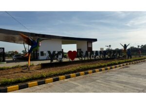 Public art display at the roadside: oversized letters with a red heart between words, flanked by a colorful hummingbird sculpture and a modern building in the background, on a paved road with a yellow-and-black curb nearby.