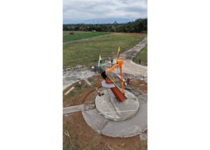 Colorful outdoor sculpture under construction on a circular concrete base in a park, with two flagpoles nearby, workers, and open grassy fields in the background.