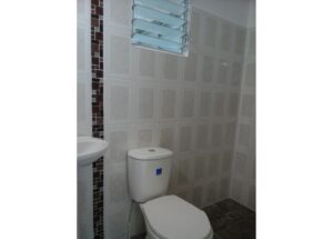 Small bathroom showing a white toilet and sink, gray tile walls with a brown mosaic vertical stripe, and a vented window above.