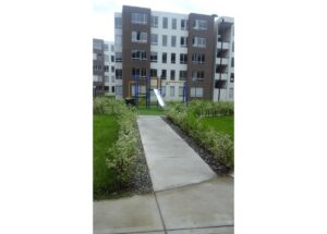 Concrete path to a children's playground with a metal slide, surrounded by trimmed hedges in a modern apartment complex.