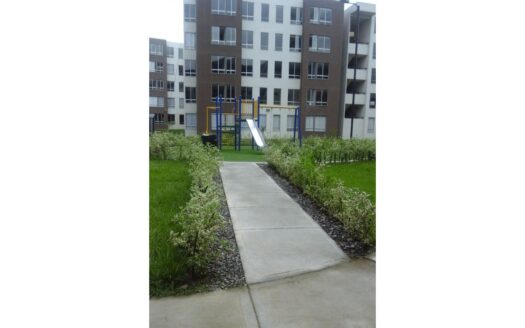 Concrete path to a children's playground with a metal slide, surrounded by trimmed hedges in a modern apartment complex.