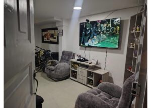 Living room basement with gray recliners, a TV mounted on the wall, and a console table; bicycles stored in the corner.