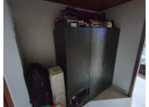 Tall black wardrobe with three doors in a corner, cluttered on top and a backpack and cardboard box leaning against it on the left side.
