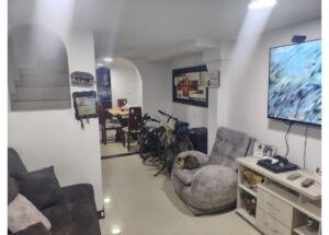 Bright basement living area with two grey couches, a TV on the wall, and bicycles parked by a dining space.