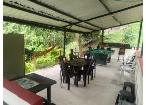 Covered outdoor recreation area with a pool table, plastic chairs, and a hammock overlooking a green hillside.