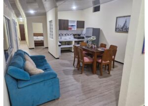 Open-plan living area with a blue sofa, wooden dining table for six, and a small kitchen in the background; a bedroom is visible down a hallway.